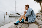 A man in a denim jacket and straw hat walks along a marina pier wearing Don't Rock The Boat blue nautical socks above black slip-on shoes. [lf]