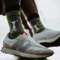 A close-up of Hang In There olive green socks featuring a tuxedo cat clinging to a branch with the phrase Hang In There, paired with cream and white New Balance sneakers. [lf]