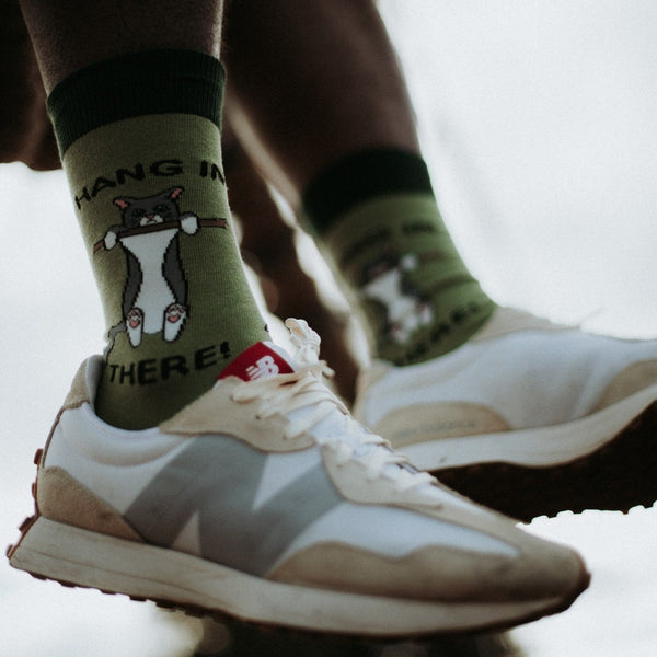 A close-up of Hang In There olive green socks featuring a tuxedo cat clinging to a branch with the phrase Hang In There, paired with cream and white New Balance sneakers. [lf]