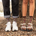 A couple's feet stand on a forest floor wearing Colorful Caps Bamboo dark brown socks with mushroom motifs, paired with white sneakers and cream platform sandals beside a basket of golden mushrooms. [lf]