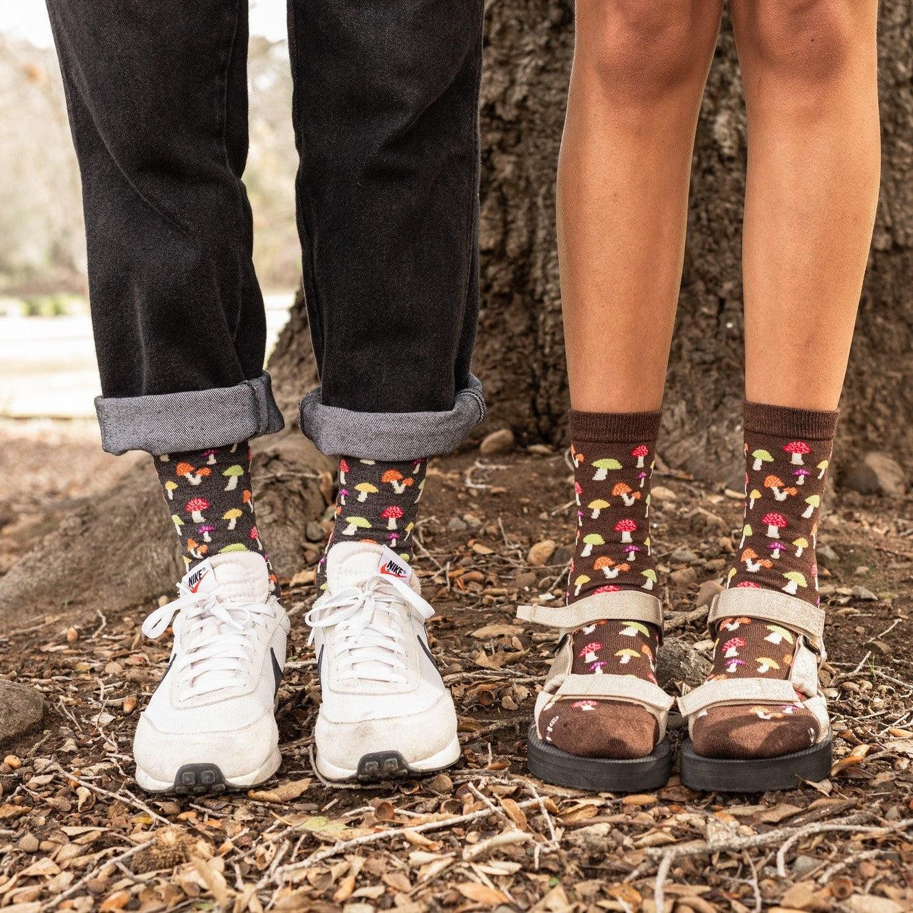 A couple's feet stand on a forest floor wearing Colorful Caps Bamboo dark brown socks with mushroom motifs, paired with white sneakers and cream platform sandals beside a basket of golden mushrooms. [lf]