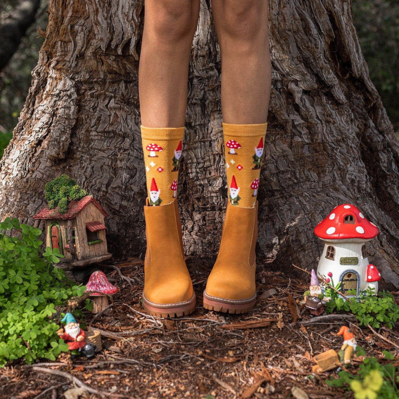 A model in yellow Gnome More Mushrooms Bamboo socks peeks out of tan rubber boots in a fairy garden beside red mushroom houses and tiny gnomes. [lf]
