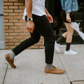Two pedestrians stride along a sunny sidewalk, the man in cream and black striped Sailor Stripe Bamboo socks above brown lace-up shoes and black skinny jeans. [lf]