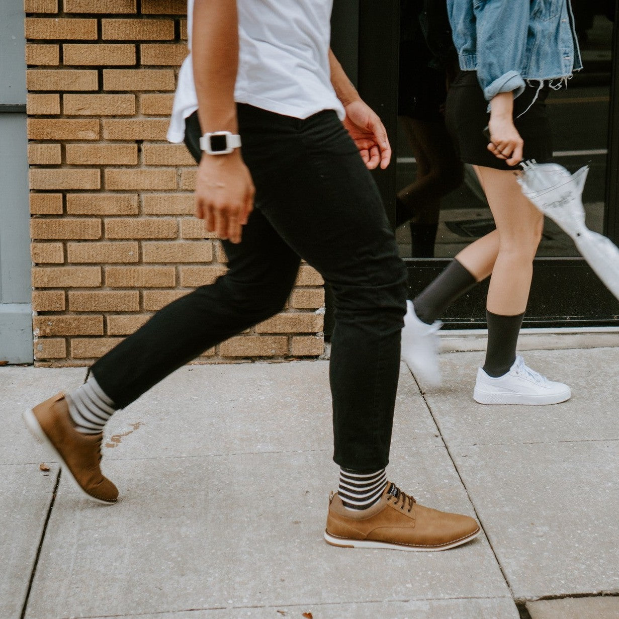 Two pedestrians stride along a sunny sidewalk, the man in cream and black striped Sailor Stripe Bamboo socks above brown lace-up shoes and black skinny jeans. [lf]