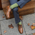 A close-up of crossed legs in grey trousers showing green and white striped Sailor Stripe Bamboo socks emerging above tan leather Oxford shoes on a leaf-covered pavement. [lf]