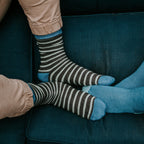 An overhead view of two people on a teal sofa showing brown and cream striped Sailor Stripe Bamboo socks beside a coordinating solid blue bamboo pair. [lf]