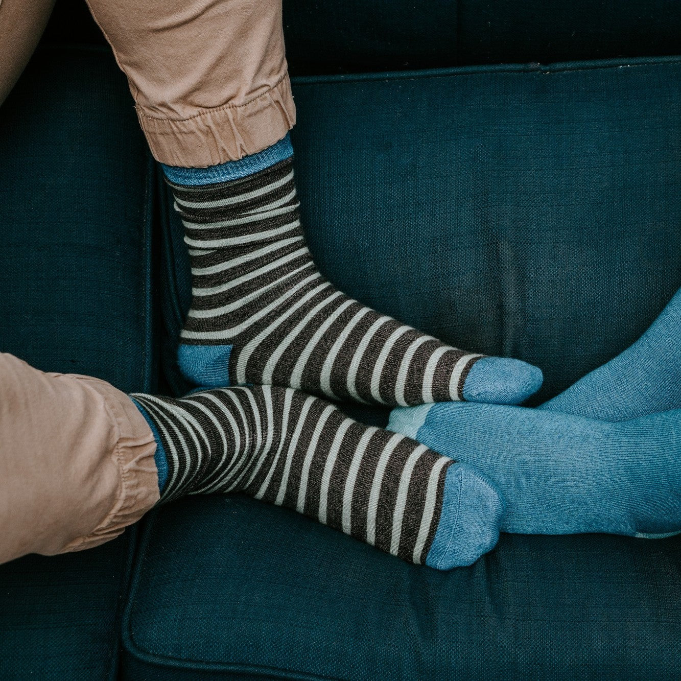 An overhead view of two people on a teal sofa showing brown and cream striped Sailor Stripe Bamboo socks beside a coordinating solid blue bamboo pair. [lf]