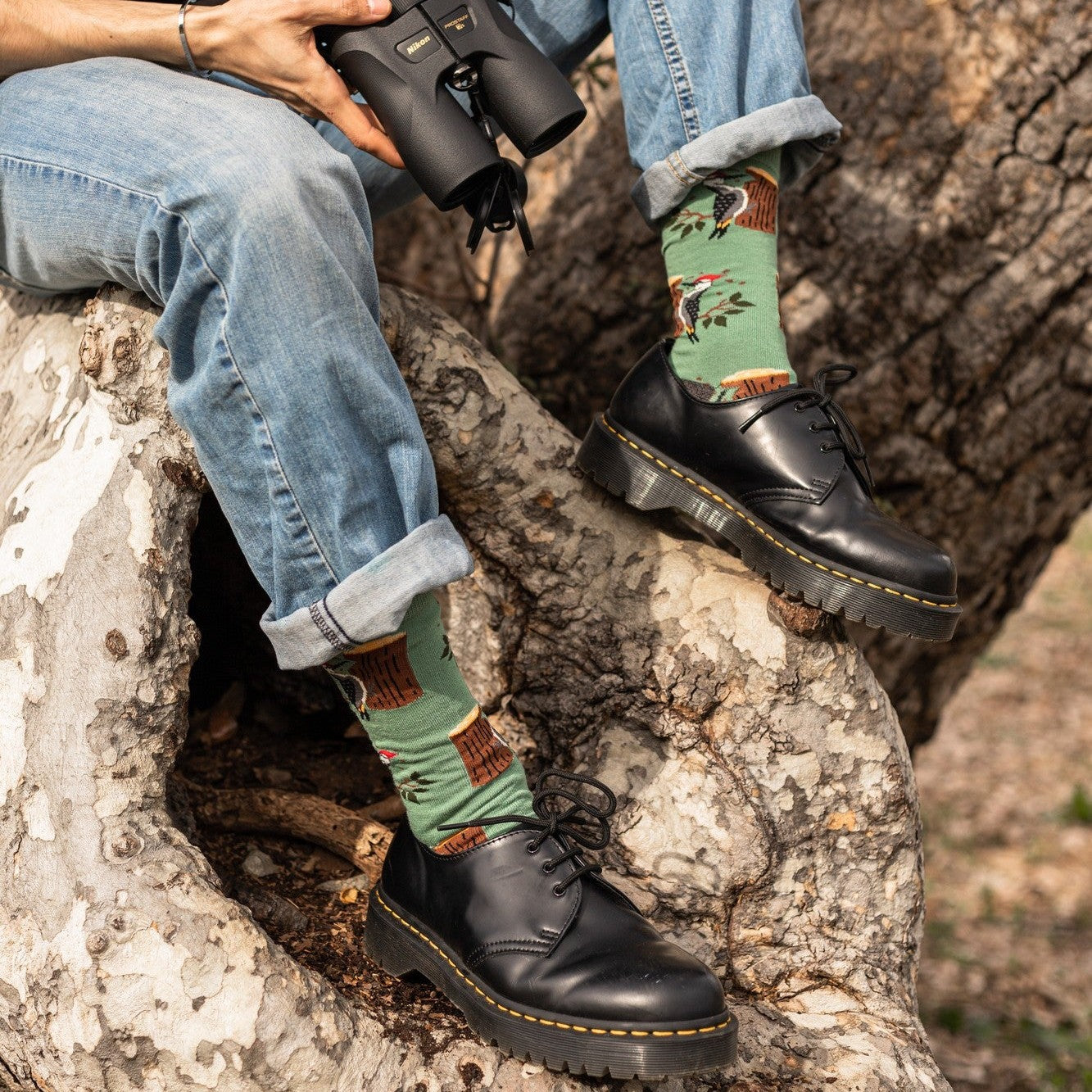 A close-up of Woodpecker Bamboo green bamboo socks featuring woodpecker motifs, paired with cuffed jeans and lace-up boots on a tree trunk. [lf]