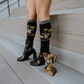 A smiling woman sits in a rattan chair in a floral mini-skirt and Goat black socks featuring tiny goat motifs, paired with shiny black heeled boots. [lf]
