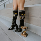 A smiling woman sits in a rattan chair in a floral mini-skirt and Goat black socks featuring tiny goat motifs, paired with shiny black heeled boots. [lf]