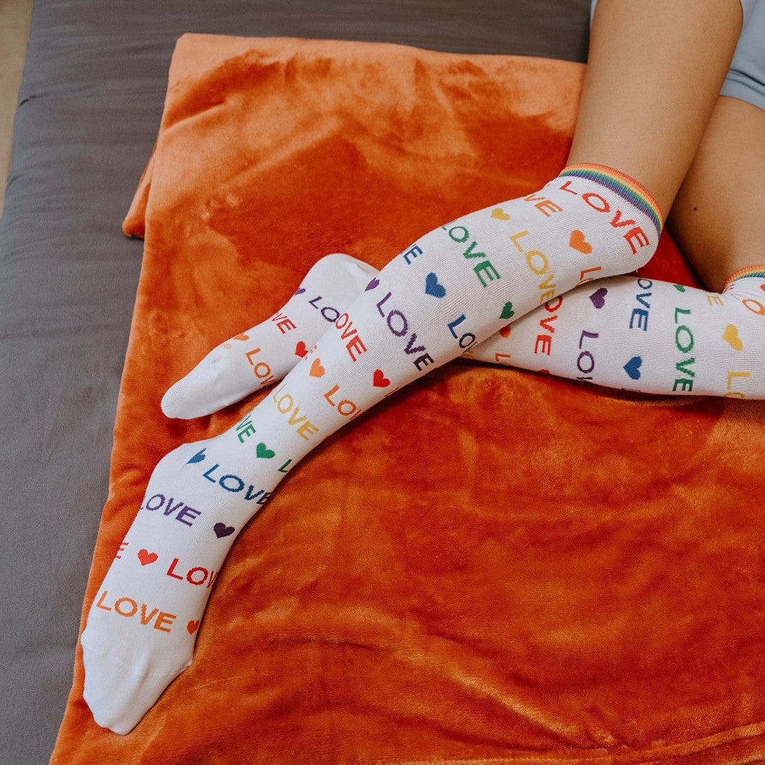 A model lounges on an orange bedspread in Love Love Love white knee-high socks with rainbow LOVE text and tiny hearts, a book open beside her. [lf]
