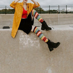 A model in a yellow jacket and red top sits on a concrete wall wearing Mexican Serape knee-high socks with rainbow stripes and chunky black boots. [lf]