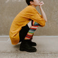 A close-up of a model's legs in Mexican Serape rainbow striped knee-high socks paired with black platform boots against a cloudy sky. [lf]