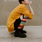 A close-up of a model's legs in Mexican Serape rainbow striped knee-high socks paired with black platform boots against a cloudy sky. [lf]