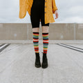 A model in a yellow jacket and red top sits on a concrete wall wearing Mexican Serape knee-high socks with rainbow stripes and chunky black boots. [lf]