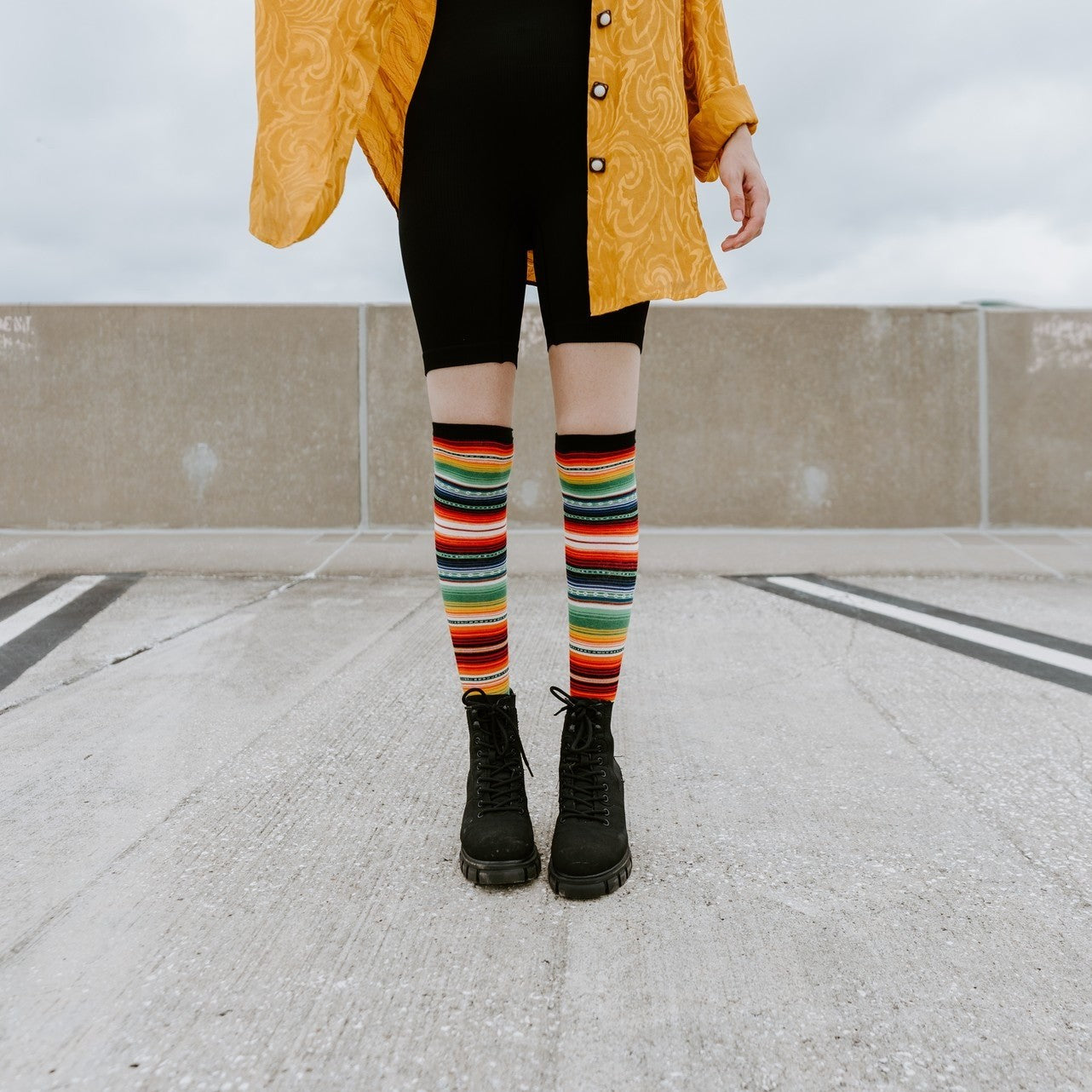 A model in a yellow jacket and red top sits on a concrete wall wearing Mexican Serape knee-high socks with rainbow stripes and chunky black boots. [lf]