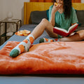 A close-up of Orange Fuzzy plush socks featuring orange fruits and green leaves, worn on a model reclined on a rust-colored blanket. [lf]