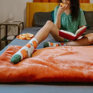 A close-up of Orange Fuzzy plush socks featuring orange fruits and green leaves, worn on a model reclined on a rust-colored blanket. [lf]