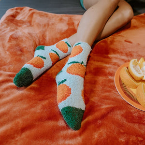 A woman reads in bed in a green dress wearing Orange Fuzzy socks with green and orange citrus prints across a fuzzy white knit. [lf]