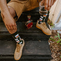A man sits on dark wooden steps wearing Whiskey Business Bamboo green socks with whiskey decanter prints, paired with tan Nike Air Force 1 sneakers, beside a crystal whiskey decanter. [lf]
