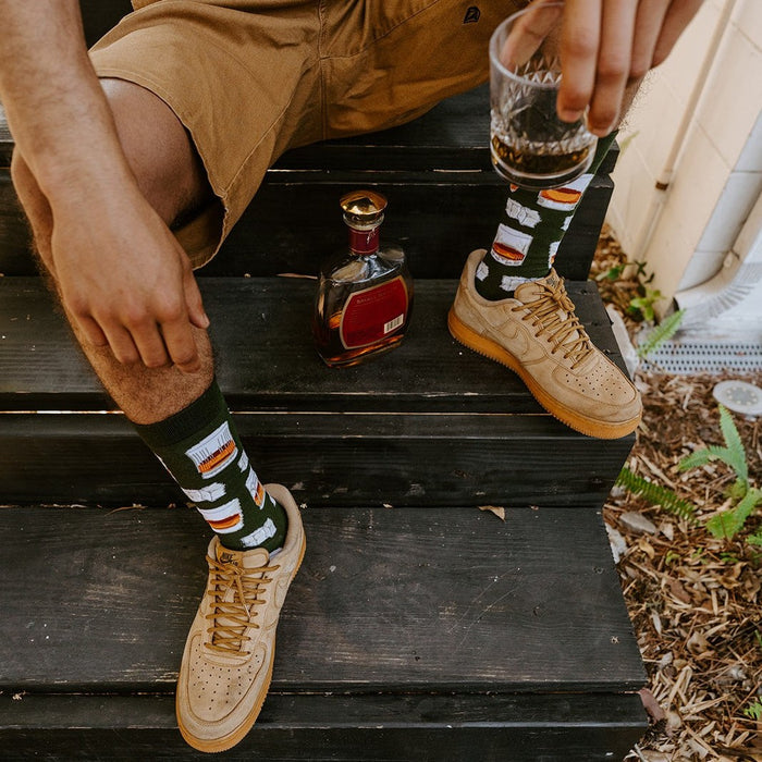 A man sits on dark wooden steps wearing Whiskey Business Bamboo green socks with whiskey decanter prints, paired with tan Nike Air Force 1 sneakers, beside a crystal whiskey decanter. [lf]