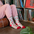 A woman in pink loungewear sits on a leather ottoman in front of a record collection, wearing Absolutely Cherry cream socks with red cherry prints and red toe and heel accents. [lf]