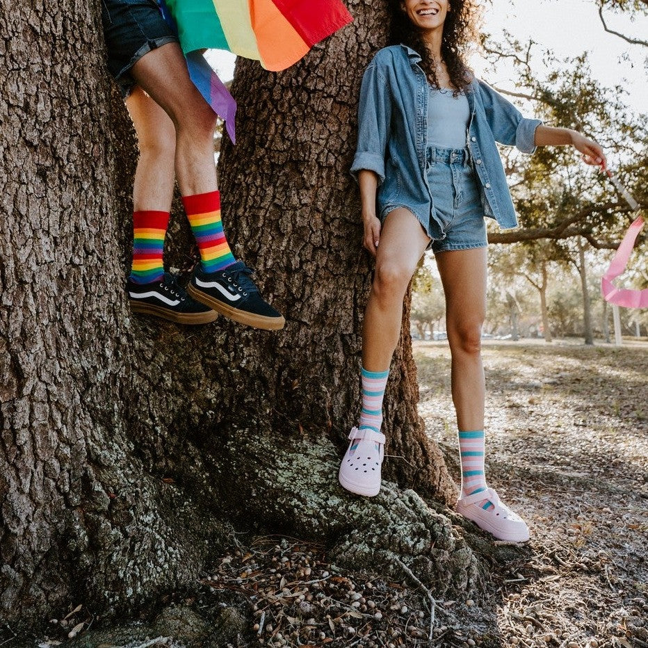 Pride Stripes rainbow socks shown on a model dancing across a grassy field with a colorful pride fan. [lf]