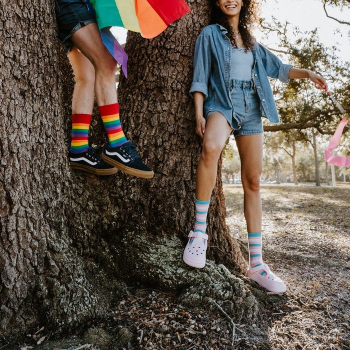 Pride Stripes rainbow socks shown on a model dancing across a grassy field with a colorful pride fan. [lf]