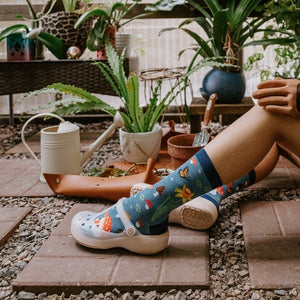 Magical Forest blue socks worn by a model tending plants in a glass greenhouse with ceramic mushroom decor. [lf]