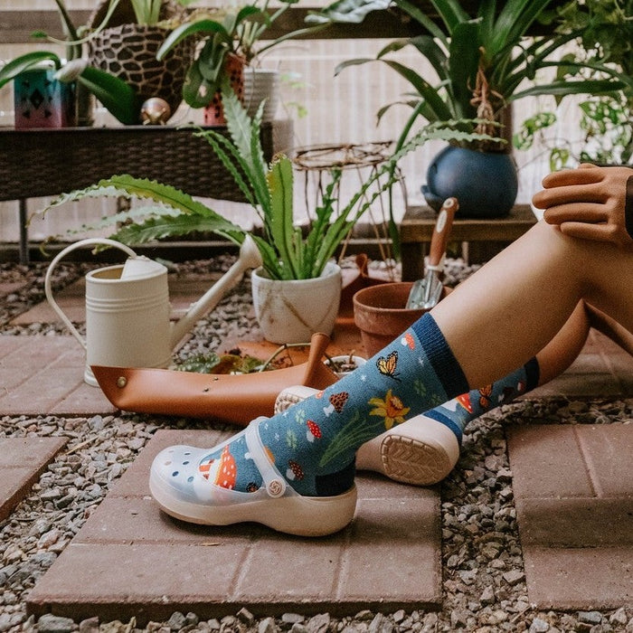 Magical Forest blue socks worn by a model tending plants in a glass greenhouse with ceramic mushroom decor. [lf]
