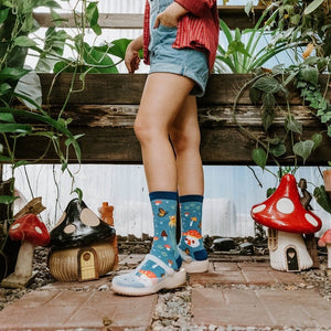 A model in red flannel sits cross-legged on a greenhouse floor wearing Magical Forest mushroom-print socks and white Crocs. [lf]