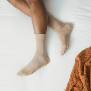 A flat-lay of Solid Bamboo socks in a soft camel hue, displayed on white bedding with a rust waffle throw beside. [lf]