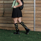 Cocktail Olives black knee-highs with martini olive motifs shown on a model leaning against a wooden fence. [lf]
