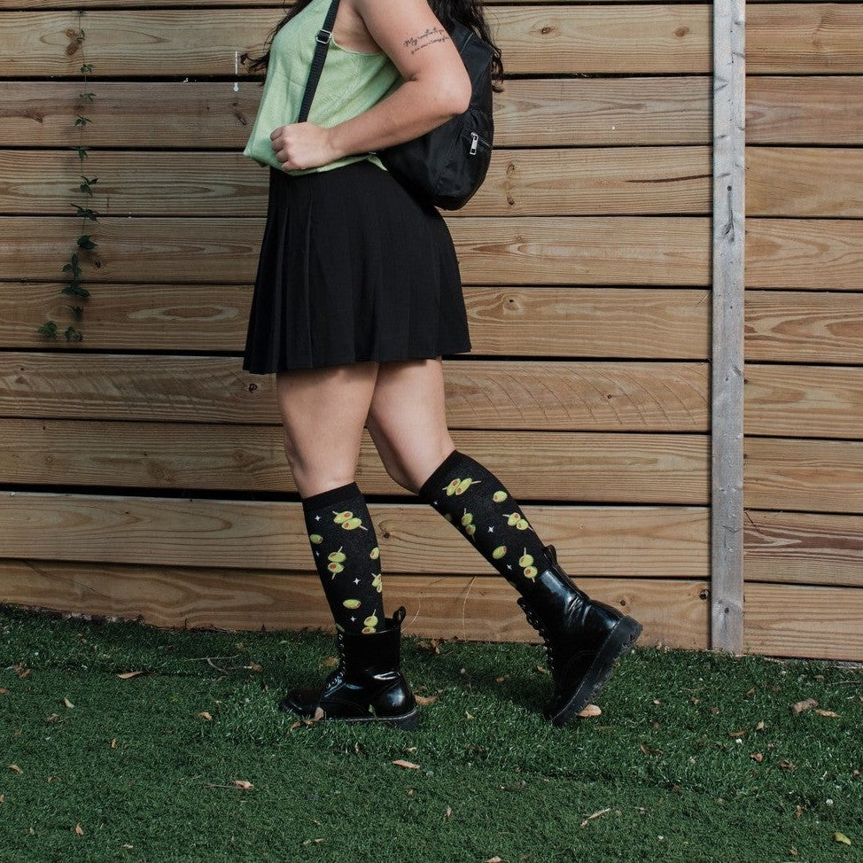 Cocktail Olives black knee-highs with martini olive motifs shown on a model leaning against a wooden fence. [lf]