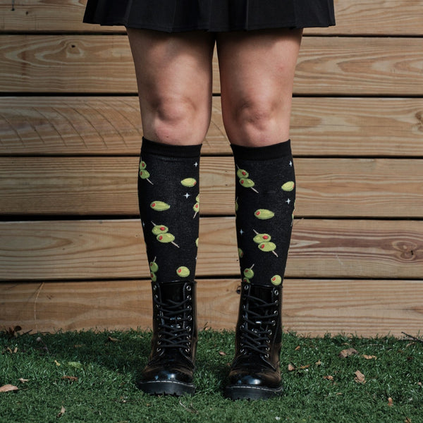 Cocktail Olives knee-high socks photographed on a model with a backpack against a fence backdrop. [lf]