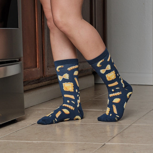 Hasta La Pasta navy socks featuring yellow pasta noodles shown on a model cooking in a sunny kitchen. [lf]