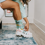 A woman in a green and white striped dress sits on a bed adjusting white platform sandals over Bird's Eye View socks featuring tropical palm and bird prints. [lf]