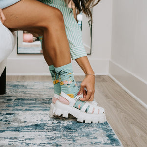 A woman in a green and white striped dress sits on a bed adjusting white platform sandals over Bird's Eye View socks featuring tropical palm and bird prints. [lf]