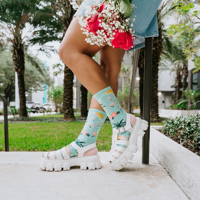 Bird's Eye View aqua socks with palm tree prints shown on a model wearing chunky white sandals on a patterned rug. [lf]