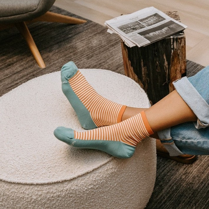 Sailor W/ Contrast Footbed socks in orange and white stripes with teal footbeds shown on a model at a modern lounge setup. [lf]