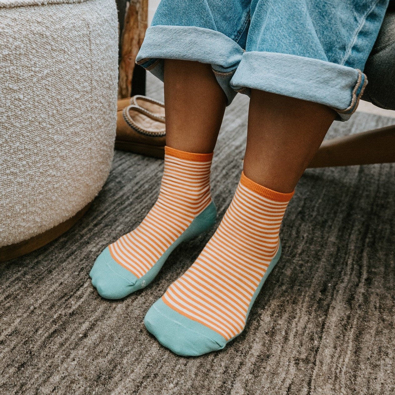 A woman works on a laptop reclining in a grey armchair, wearing Sailor W/ Contrast Footbed striped socks with orange footbeds and tan moccasins beside the ottoman. [lf]