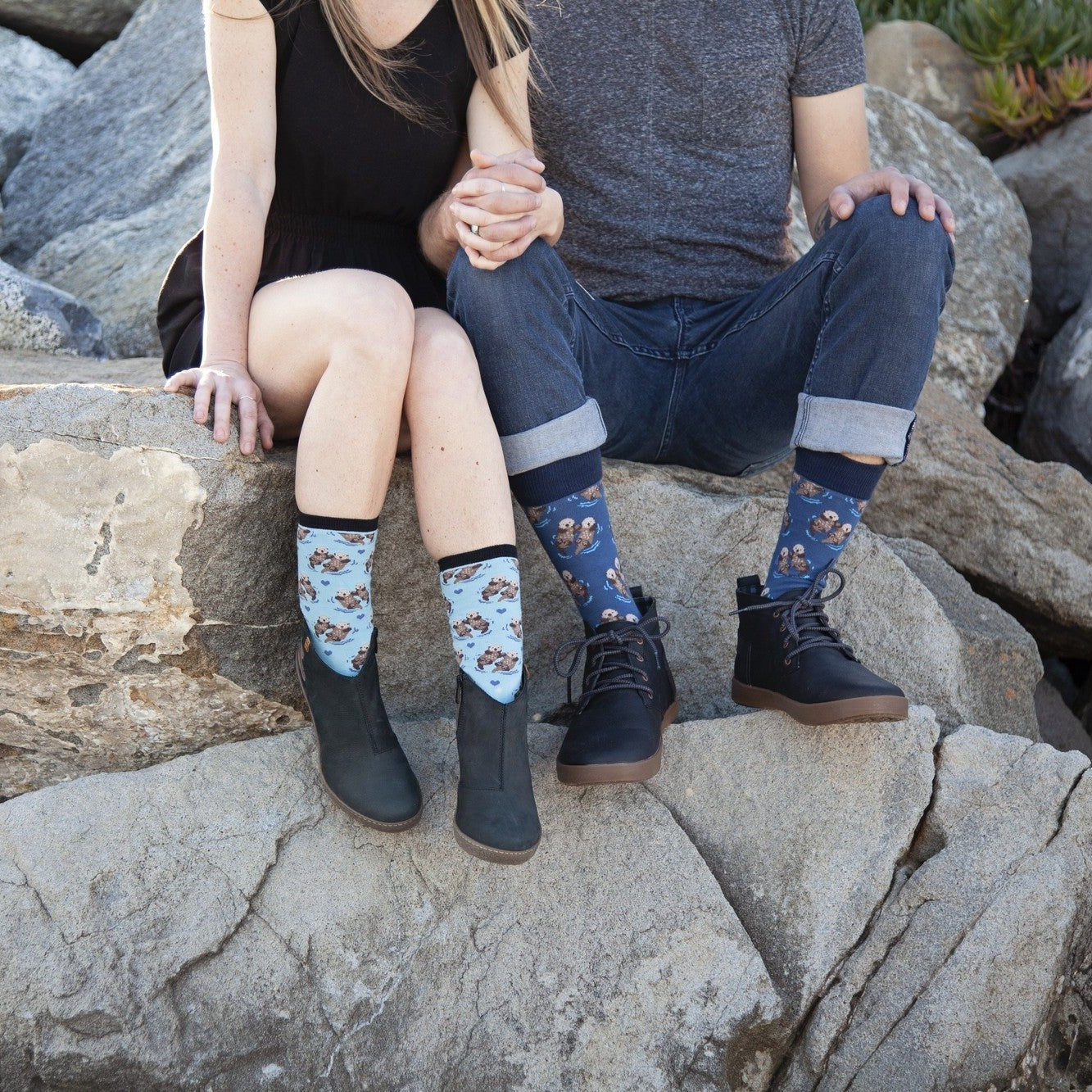 A couple sits side by side on sun-warmed beach rocks holding hands, the woman wearing light Blue Significant Otter socks with floating otter prints and the man wearing Black Significant Otter socks tucked into dark boots. [lf]