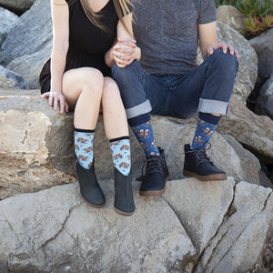 A couple sits side by side on sun-warmed beach rocks holding hands, the woman wearing light Blue Significant Otter socks with floating otter prints and the man wearing Black Significant Otter socks tucked into dark boots. [lf]