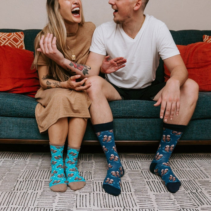 A couple shares a laugh on a teal couch with red and orange pillows, both showing off their matching Significant Otter socks in light Blue and dark Blue otter prints. [lf]