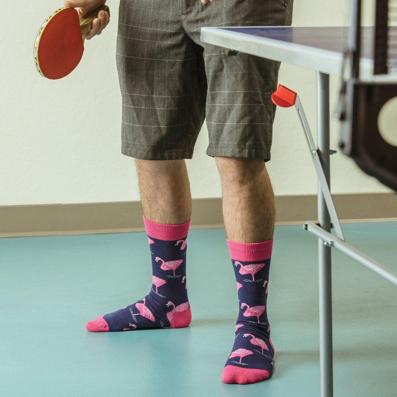 A ping-pong player in a yellow tee and gray plaid shorts holds a red paddle and orange ball beside a folding table, his Flamingo socks in NAVY scattered with pink flamingos and finished with hot-pink cuffs and toes. [lf]
