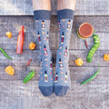 A pair of feet on weathered wood planks framed by scattered habanero, jalapeno, and serrano peppers plus bottles of Tabasco, the wearer in denim-blue Hot In Here socks printed with hot sauce bottles and chili flakes. [lf]