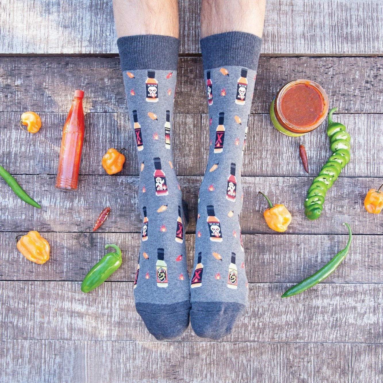 A pair of feet on weathered wood planks framed by scattered habanero, jalapeno, and serrano peppers plus bottles of Tabasco, the wearer in denim-blue Hot In Here socks printed with hot sauce bottles and chili flakes. [lf]