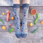 A pair of feet on weathered wood planks framed by scattered habanero, jalapeno, and serrano peppers plus bottles of Tabasco, the wearer in denim-blue Hot In Here socks printed with hot sauce bottles and chili flakes. [lf]