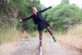 Joyful woman balances on a rusted rail with arms outstretched, black dress fluttering, her boots topped with Spooky Season Sweater socks in Black and Orange showing colorful folk-knit cuffs. [lf]