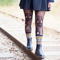 Cropped view of skull tights, a black skater dress, and chunky Chelsea boots resting on rusted tracks, the boot tops trimmed by Spooky Season Sweater socks in Black and Orange with intricate Halloween Fair Isle bands. [lf]